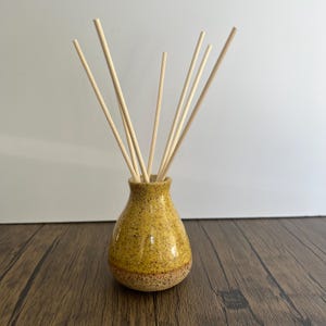 May include: A small, round, yellow ceramic diffuser vessel with a speckled texture and a narrow neck. Several thin, light-coloured wooden reed sticks are inserted into the opening, extending upwards and outwards from the centre of the container.