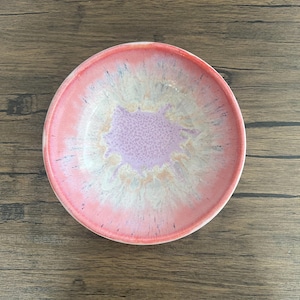May include: A round ceramic bowl with a pink rim and a centre that fades from lavender to white. The bowl's interior has a speckled design, and the exterior is a solid pink. The bowl is set on a wooden surface.