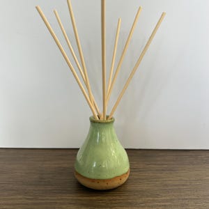 May include: A ceramic reed diffuser featuring a light green glazed upper section and a natural terracotta base. The vessel has a rounded, tapered neck and contains several thin, light-coloured wooden reeds extending outward from the opening.