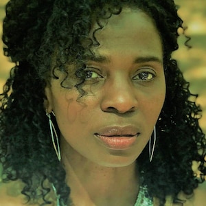 May include: A woman with dark curly hair wearing a silver necklace and earrings. She is looking at the camera with a neutral expression.