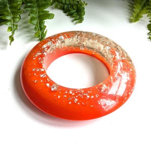 May include: A circular bangle bracelet made of smooth, vibrant orange resin. The surface is embedded with numerous small, reflective silver-coloured metallic flakes that are distributed throughout the translucent and opaque material.