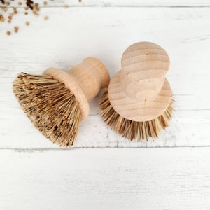 May include: Two wooden dish brushes with natural bristles. The brushes are on a white wooden surface.