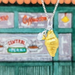 May include: A silver necklace with a gold croissant charm and a small silver coffee cup charm. The necklace is in front of a green and orange building with the words "Central Perk" on it.