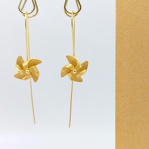 Puede incluir: Pendientes dorados con diseño de molinillo. Los pendientes tienen un alambre largo y fino que se conecta a un pequeño molinillo texturizado. Los molinillos tienen un pequeño centro redondo. Los pendientes cuelgan de un alambre curvado.