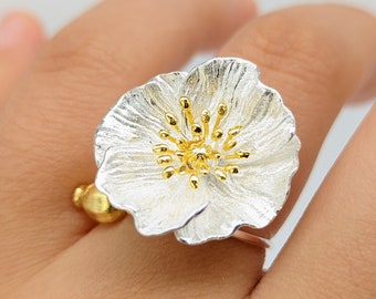 May include: A silver ring with a white flower design. The center of the flower is gold and has a textured design. The ring is on a finger.