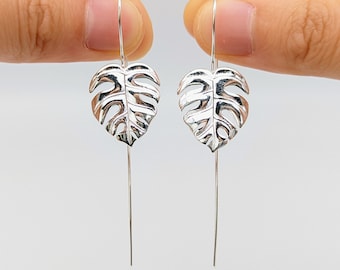 May include: Silver dangle earrings featuring a delicate monstera leaf design. The earrings are long and thin, with a simple hook closure.