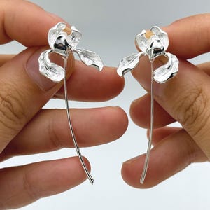 Pode incluir: Um par de brincos prateados em forma de flores de íris. Cada brinco apresenta uma cabeça de flor detalhada com pétalas delicadas e uma haste longa e curva. Os brincos são apresentados em um fundo neutro.