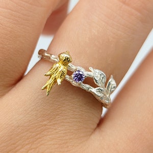 May include: A silver ring with a delicate branch design and a purple gemstone. A small gold bird is perched on the branch.