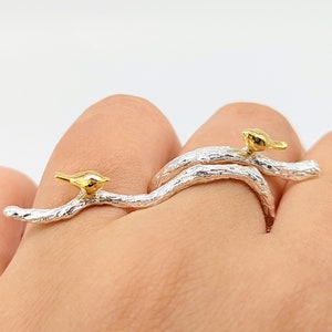 Op de afbeelding: Een zilveren ring met twee gouden vogels die op takken zitten. De ring is ontworpen om op twee vingers te worden gedragen.