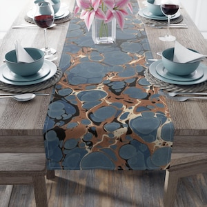 Op de afbeelding: Een blauwe en bruine marmeren tafelrunner met een draaiend patroon. De runner ligt op een houten tafel met couverts aan beide kanten.