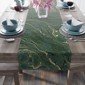 Op de afbeelding: Een groene en crèmekleurige tafelrunner met een marmerpatroon. De runner ligt op een houten tafel met twee couverts aan elke kant.