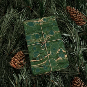 May include: A green and gold marbled gift box tied with twine, nestled amongst pine needles and pine cones.