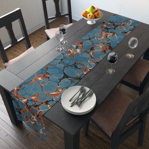 Op de afbeelding: Een blauwe en oranje marmeren tafelkleed met een draaiend patroon. De loper ligt op een donkerhouten tafel met twee stoelen en een glas wijn.