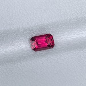 Puede incluir: Una gema de color rosa intenso y profundo tallada en forma de esmeralda rectangular. La piedra presenta bordes facetados precisos que reflejan la luz, mostrando un color rico y saturado en toda su estructura cristalina transparente y pulida.