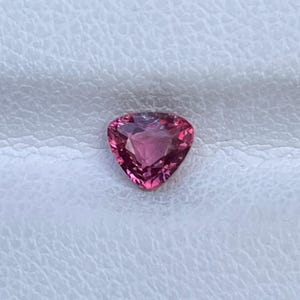 Puede incluir: Una piedra preciosa facetada de forma triangular con un tono rojo rosado vibrante. La piedra presenta un corte brillante con múltiples facetas geométricas que reflejan la luz, creando una apariencia clara y brillante en su superficie.