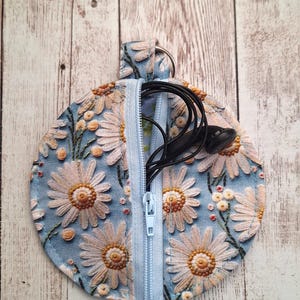 May include: A circular, light blue pouch with a daisy floral pattern and a zip. The pouch is open, showing black earbuds inside. It has a small loop and a silver ring at the top.