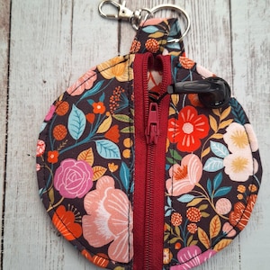 May include: A circular floral pouch with a red zip and a silver keyring clip. The pouch has a dark background with pink, orange, and blue flowers. A black earbud holder is attached to the zip.