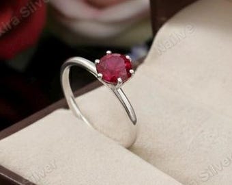 May include: A silver ring with a round, red gemstone, displayed in a brown jewellery box with a cream interior. The background features red and pink roses, creating a romantic setting.