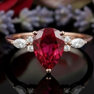 Könnte beinhalten: Roségoldener Ring mit einem birnenförmigen, tiefroten Edelstein und kleineren, klaren Steinen. Der Ring wird vor einem Hintergrund aus Rosen und Lavendel präsentiert, was eine luxuriöse und romantische Ästhetik schafft.