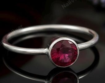 May include: A silver ring featuring a round, deep red gemstone. The ring has a thin, polished band, and the gemstone is set in a bezel. The ring is shown on a reflective black surface, with soft background lighting.
