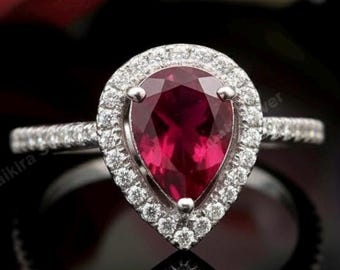 Peut inclure: Bague en argent ornée d'un rubis en forme de poire, entouré d'un halo de petits diamants. La bague est mise en valeur sur un fond flou de roses rouges, roses et violettes. Le reflet de la bague est visible sur une surface noire.
