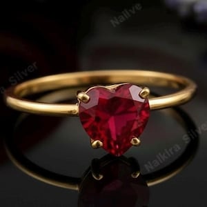 以下が含まれることがあります： ハート型の深紅色の宝石をあしらったゴールドリング。リングのバンドは細く磨かれており、宝石は4つの爪で固定されています。リングは暗い背景にセットされており、その反射性を際立たせています。