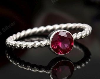 May include: A silver ring featuring a twisted band design, set with a prominent, circular, deep red gemstone. The ring is displayed against a dark backdrop, with its reflection visible.