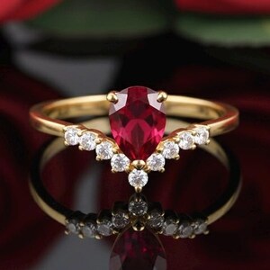 May include: A gold ring with a pear-shaped, deep red gemstone as the centerpiece. The ring features a V-shaped band adorned with small, round, white gemstones. The ring is set against a dark background with red rose petals.