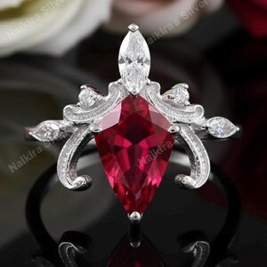 May include: A silver ring with a large, red, kite-shaped gemstone as the focal point. The ring features a decorative, ornate design with smaller white gemstones and a marquise-cut stone at the top. The ring is set against a dark background.