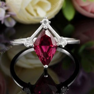 May include: A silver ring with a large, faceted, ruby-red gemstone in a kite shape. The ring has a delicate design with small, clear accent stones and a reflective surface.