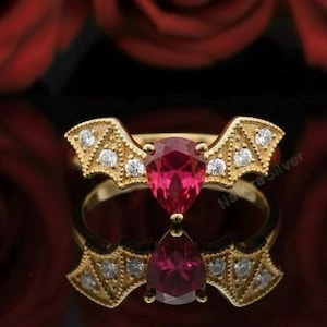 May include: A gold ring with a pear-shaped, ruby-colored gemstone at its center. The ring features a decorative wing-like design with small, clear accent stones. The ring is displayed on a reflective surface with red roses in the background.