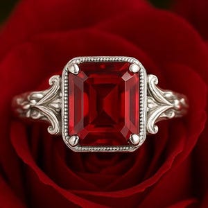 Peut inclure: Une bague en argent ornée d'une grande pierre rouge foncé de forme rectangulaire. La bague présente un motif filigrané orné sur le corps et autour de la pierre précieuse. La bague est posée sur une rose rouge, créant un contraste saisissant.