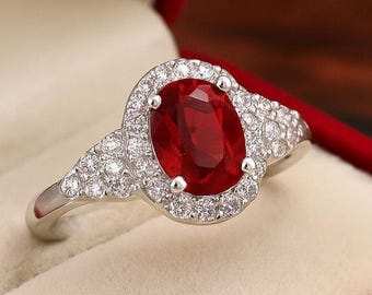 Könnte beinhalten: Ein silberner Ring mit einem großen, oval geschliffenen Rubin, umgeben von einem Kranz aus kleineren, klaren Edelsteinen. Der Ring wird in einer roten Samtbox präsentiert. Das Design des Rings umfasst zusätzliche Edelsteine ​​am Band.
