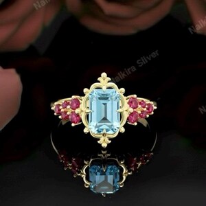 May include: A gold ring featuring an emerald-cut, light blue gemstone as the centerpiece, surrounded by smaller, round, red gemstones. The ring has an ornate, filigree design and is set against a dark background with a reflection.