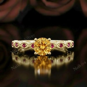 May include: A gold ring with a large, round, yellow gemstone as the centerpiece. The band is adorned with small, red gemstones and intricate detailing. The ring is set against a dark background with blurred red rose accents, creating a luxurious feel.