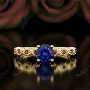 May include: A gold ring with a large, round, blue gemstone at the center. The band is detailed with small red gemstones and decorative patterns. The background features blurred red roses.