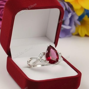 Op de afbeelding: Een zilveren ring met een peervormige, dieprode edelsteen, gezet in een decoratieve band met kleine, heldere accentstenen. De ring wordt gepresenteerd in een open, rode fluwelen ringdoos, tegen een bloemenachtergrond.