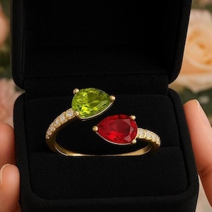 Op de afbeelding: Een gouden ring met twee peervormige edelstenen, een groene en een rode, gepresenteerd in een zwarte sieradendoos. De ring is versierd met kleine, heldere stenen langs de band. De achtergrond toont onscherpe bloemen.