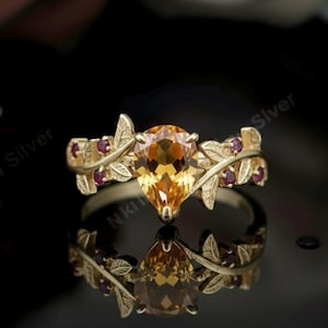 May include: A gold-toned ring featuring a pear-shaped, amber-colored gemstone as the centerpiece. The ring is adorned with leaf and berry accents, with smaller red gemstones set along the band. The ring is reflected in a dark, glossy surface.