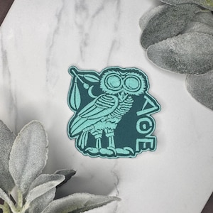 May include: An embroidered patch featuring a teal owl with large circular eyes, perched on a branch with a single leaf. The design includes a small crescent moon above the owl and the vertical text "AOE" in a stylized font on the right side.