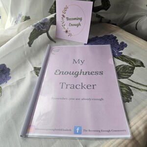 May include: A light purple "My Enoughness Tracker" journal with the text "Remember...you are already enough". A small card with the text "Becoming Enough" is also visible. The journal and card are on a floral patterned surface.