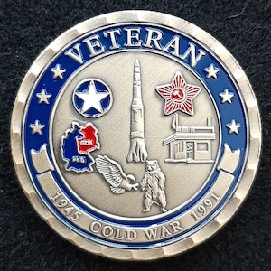May include: A silver coin with a blue border and the word "Veteran" in blue lettering. The coin features a red star with a hammer and sickle, a red and white map of Germany, a rocket, a building, a star, a bear, and a bird. The coin also has the text "1945 Cold War 1991".