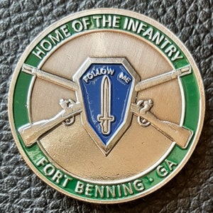 May include: A silver challenge coin with a green border. The coin features a crossed rifle design, a blue shield with a sword and the words "Follow Me", and the text "Home of the Infantry Fort Benning, GA".