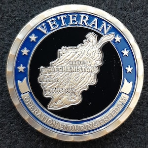 May include: A silver coin with a blue border and a black center. The coin features a map of Afghanistan with the words "Veteran" and "Operation Enduring Freedom" around the edge.