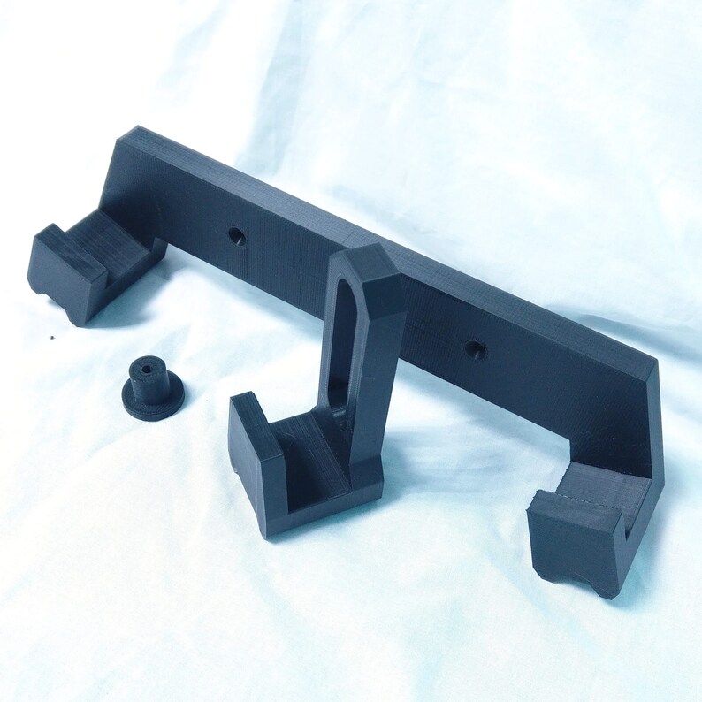 May include: A black 3D printed wall mount with two hooks. The mount has a rectangular base with two side arms that extend out and up. The hooks are located at the end of each arm.