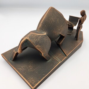 May include: A bronze sculpture of a bull with a person standing next to it. The bull is in a charging pose with its head lowered and its horns pointed forward. The person is standing upright with their arms at their sides. The sculpture is set on a rectangular base.