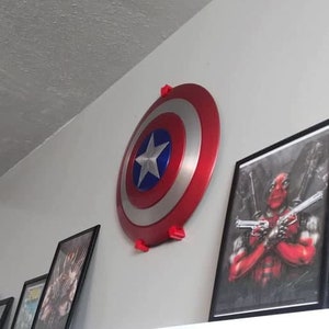 May include: A red, white, and blue Captain America shield with a five-pointed star in the center is mounted on a wall. The shield is made of metal and has a raised edge. It is hanging from two red plastic hooks.