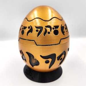 May include: A gold egg-shaped object with black markings. The egg is sitting on a black base.