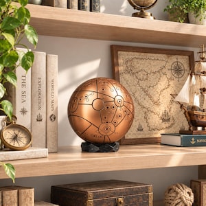 May include: A spherical copper-colored decorative object featuring intricate black etched lines and circular patterns, resting on a black faceted base. The surface displays a complex network of interconnected geometric shapes and small dots.