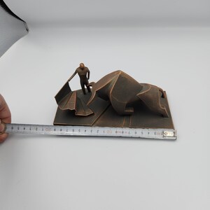 May include: A bronze-colored sculpture of a bullfighter facing a bull. The bull is lying down and the bullfighter is holding a cape. The sculpture is on a rectangular base.